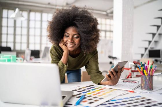 Woman graphic designer in her office looking at laptop and holding a smartphone